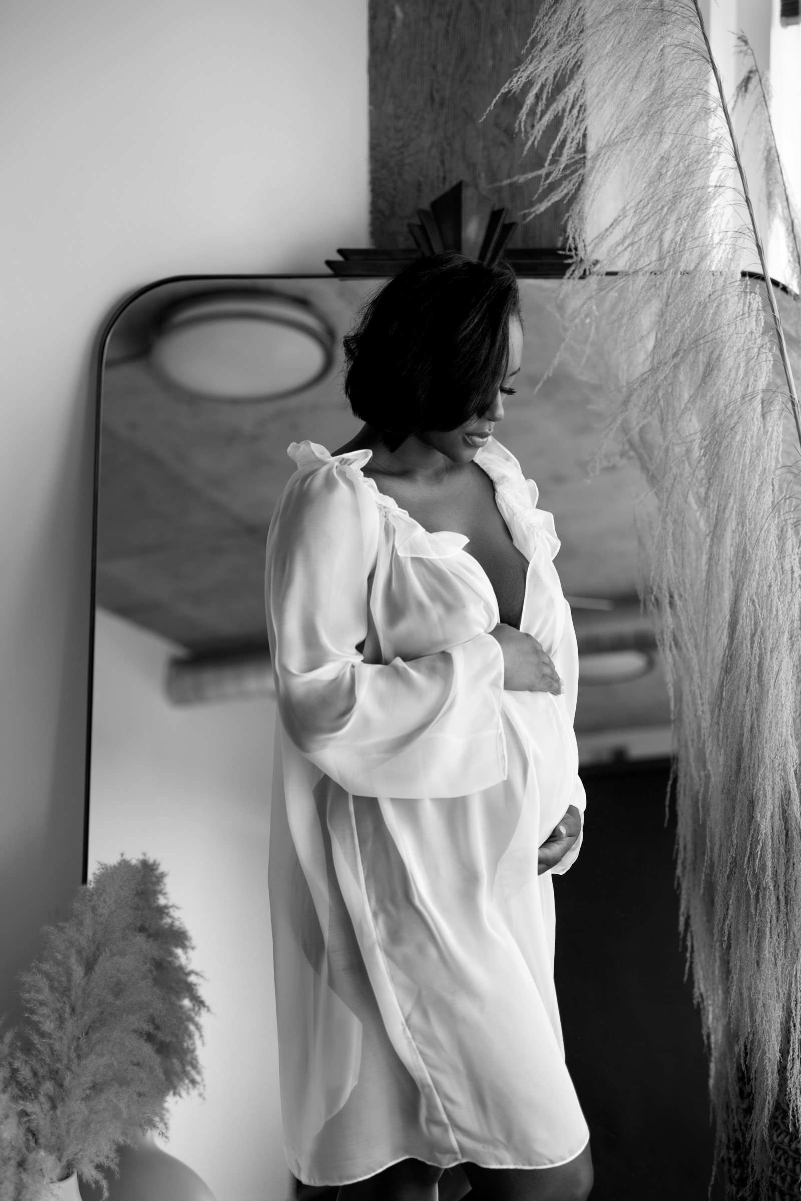 Pregnant woman wearing a white robe beside a loft-studio window and mirror