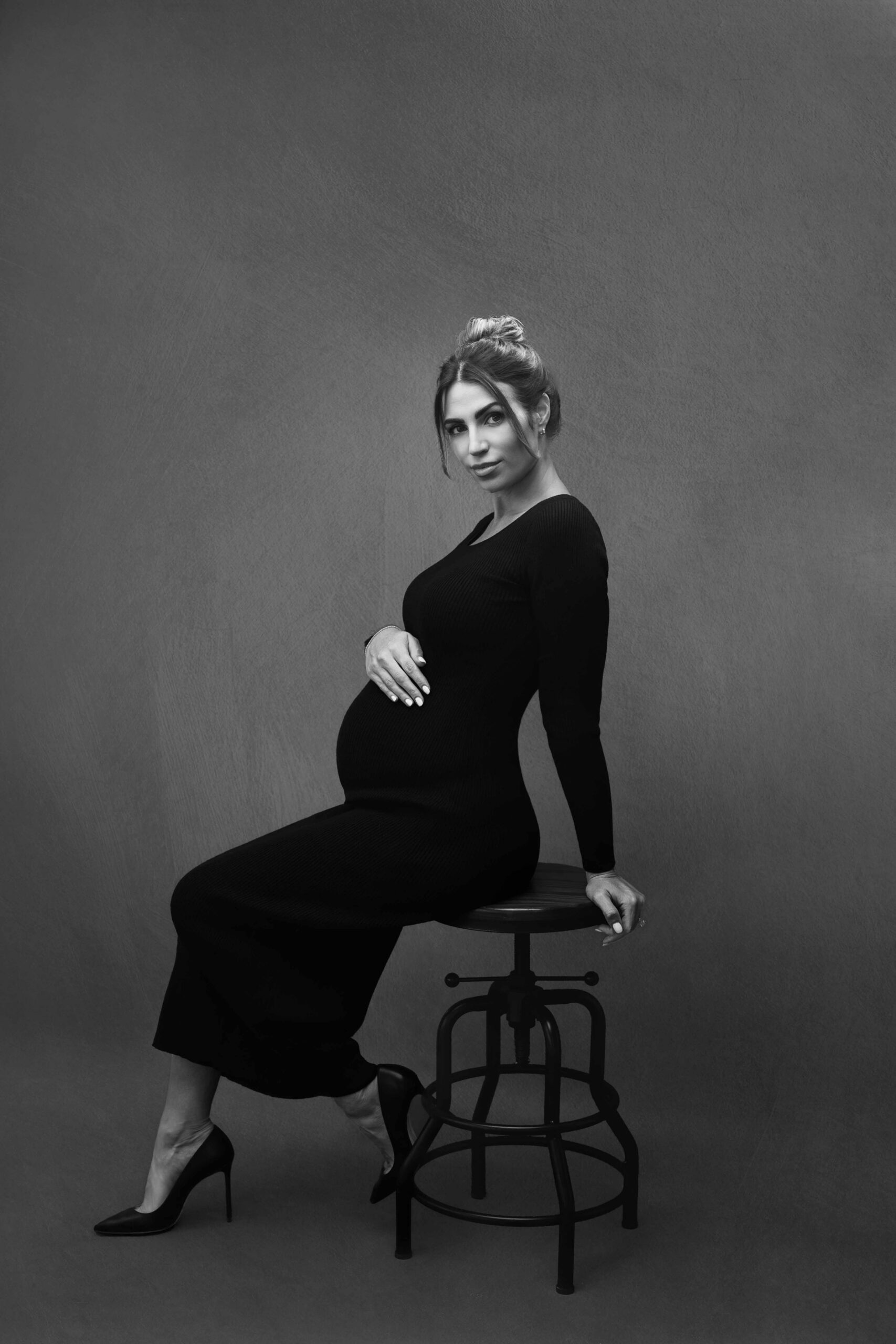 Pregnant woman in a fitted black dress seated on a studio stool