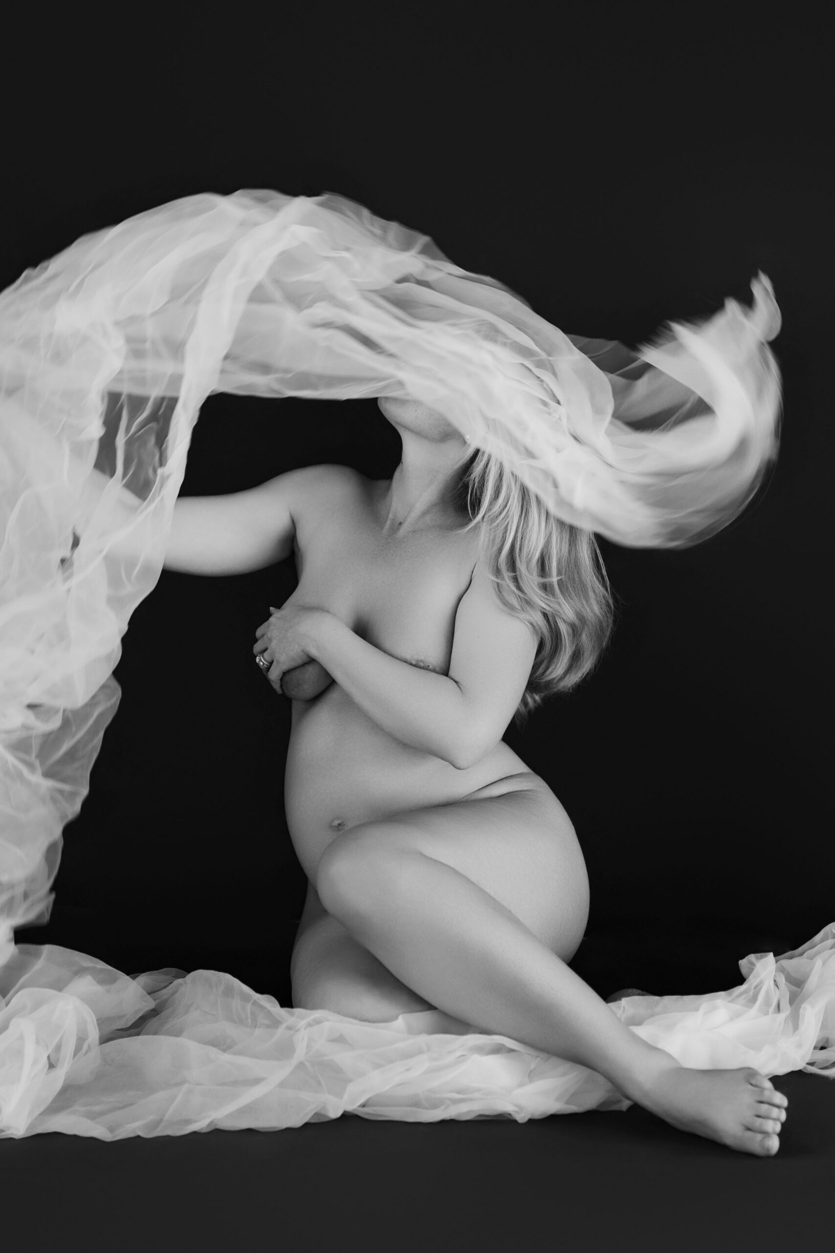 Pregnant woman seated among flowing white tulle against a dark studio backdrop