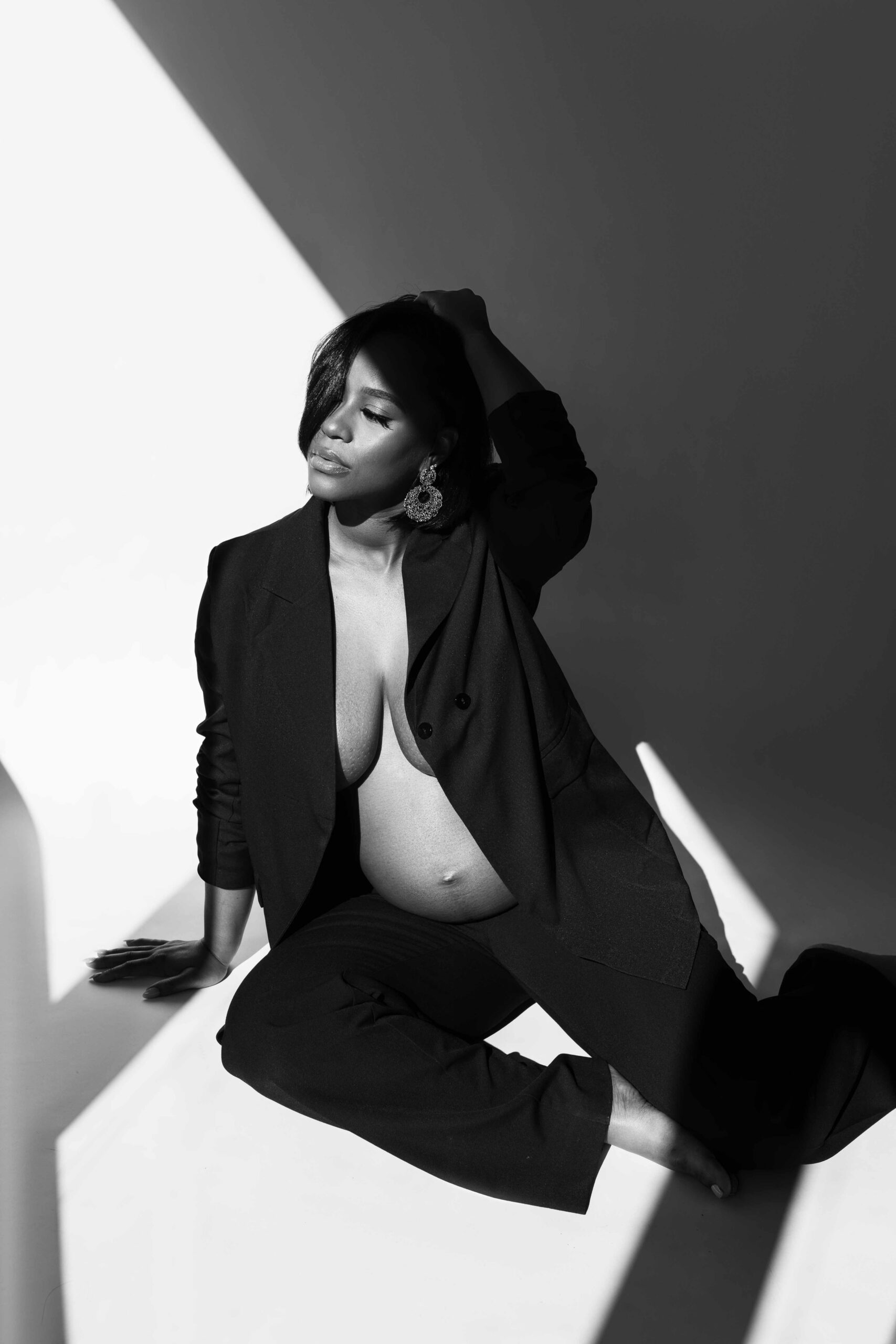 Pregnant woman seated in a black blazer and trousers under geometric studio light