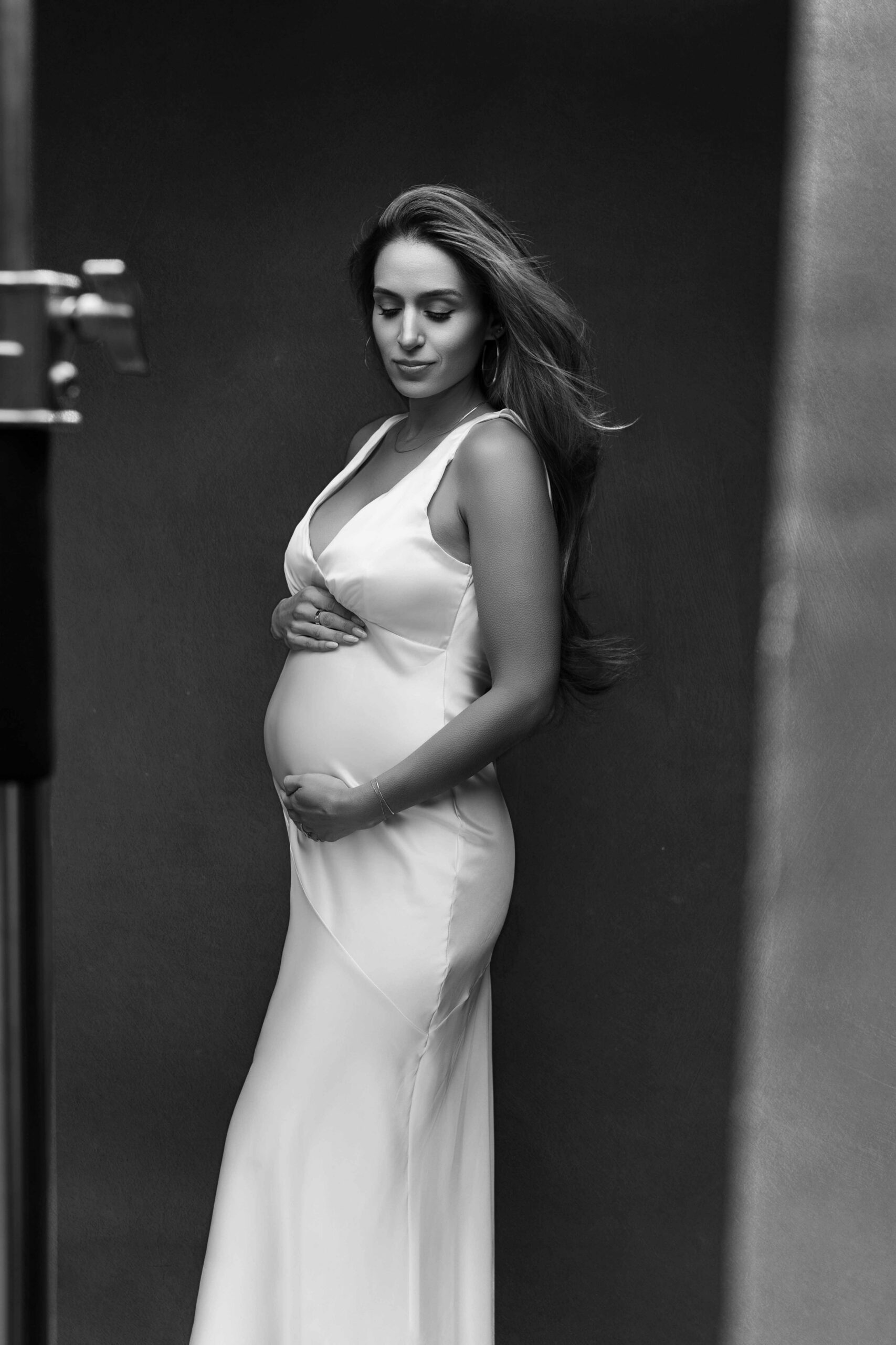 Pregnant woman in a white satin dress holding her belly in soft studio light