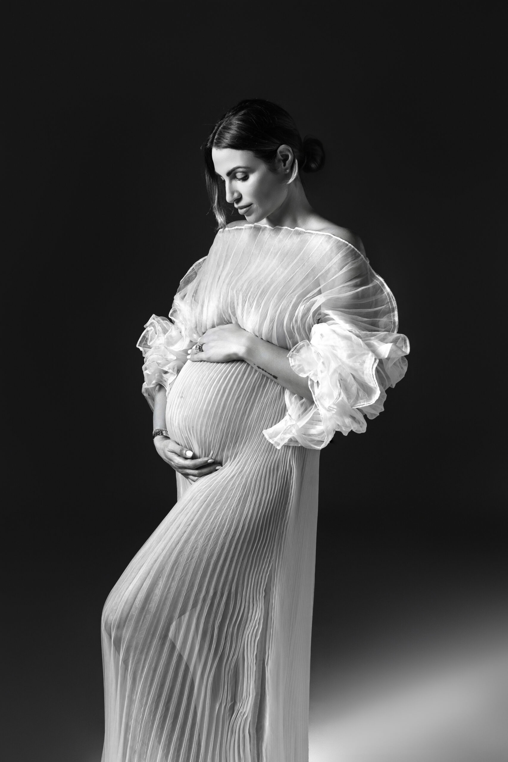 Pregnant woman wearing a pleated white gown with ruffled sleeves against a dark backdrop