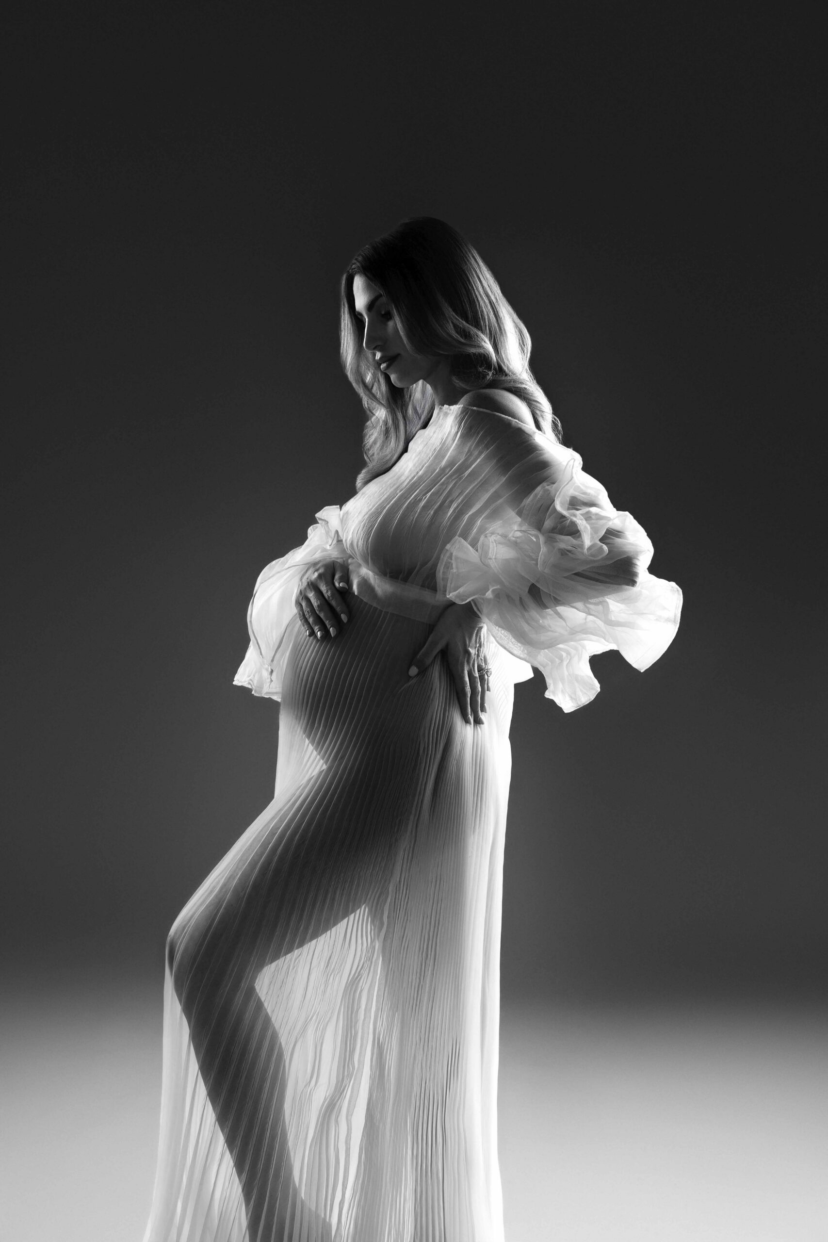Pregnant woman wearing a sheer pleated gown with ruffled sleeves in studio light