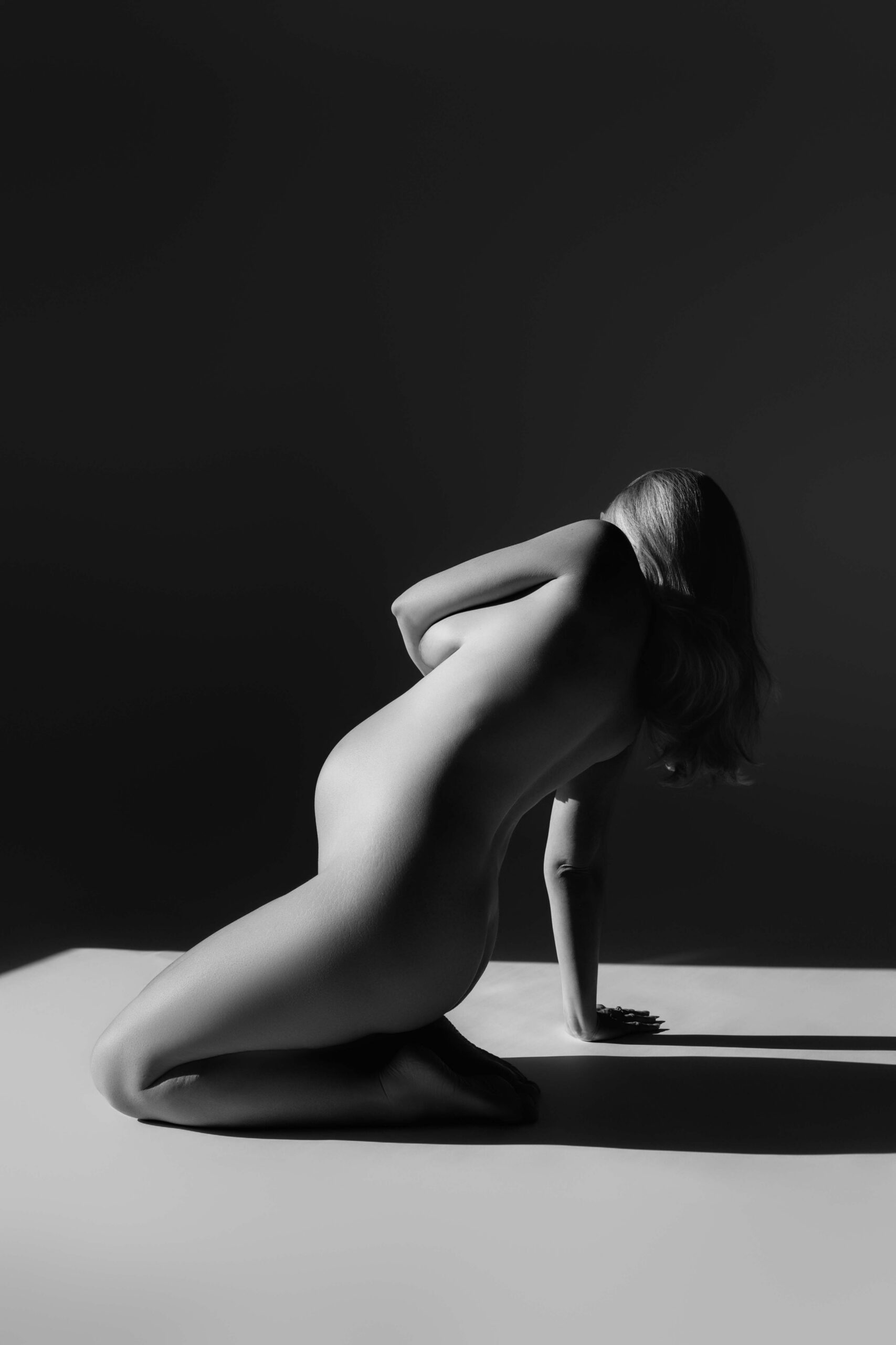 Pregnant woman kneeling in profile with curved posture and dramatic black-and-white studio light