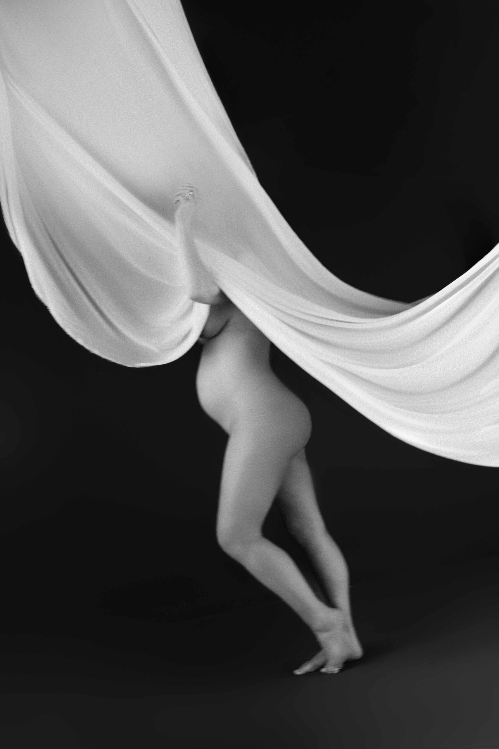 Pregnant woman partially veiled by flowing white fabric against a dark studio backdrop
